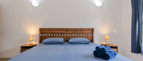 Una cama bien hecha con una colcha azul estampada, dos almohadas y toallas enrolladas a un lado. Hay dos lámparas de noche con pantallas estampadas.