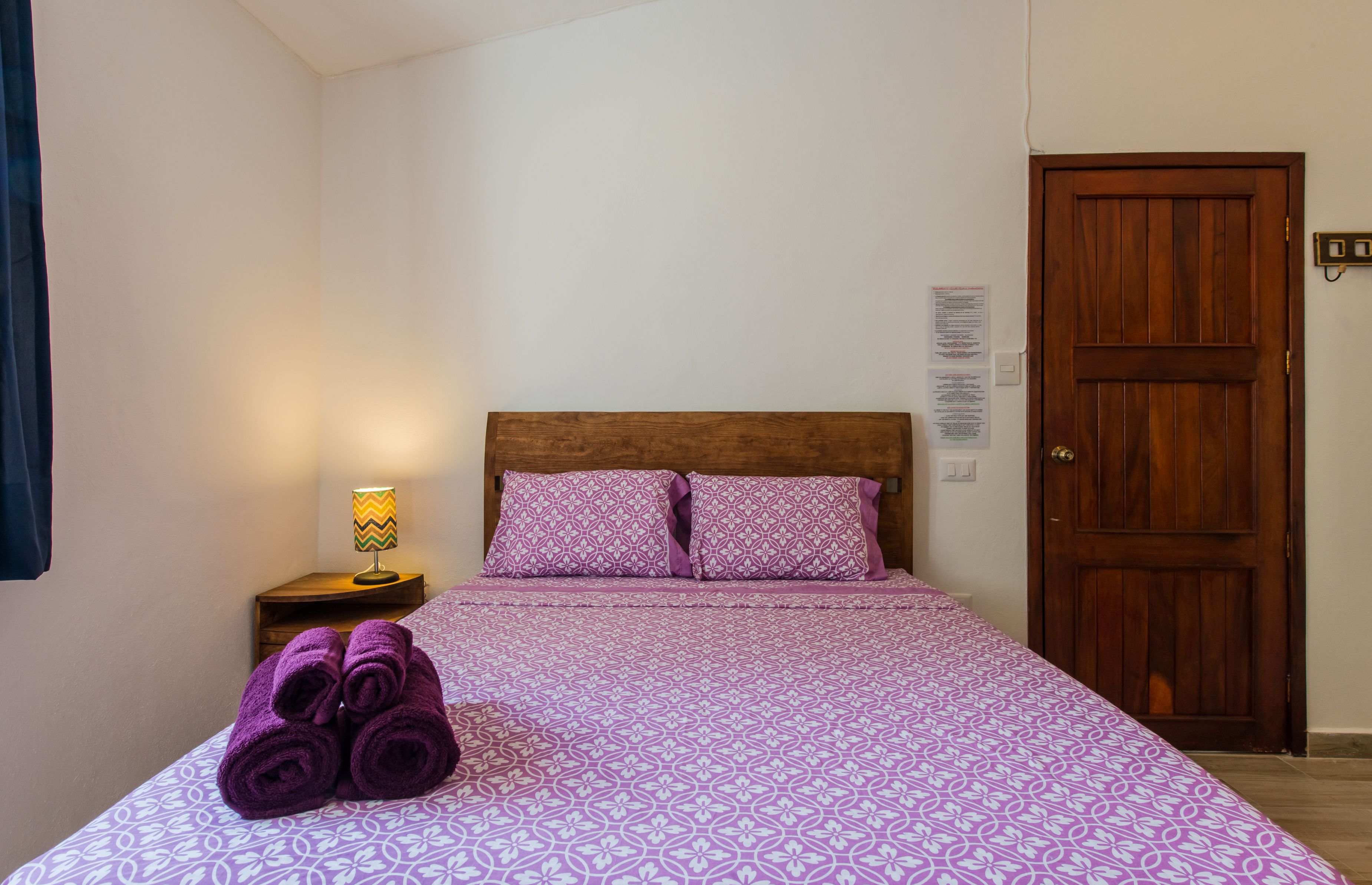 A neatly made bed with purple bedding and pillows, a wooden headboard, a nightstand with a lamp, and a wooden door.