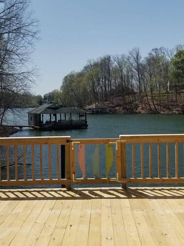A wooden deck with a railing, a covered structure by the water, and trees in the background.