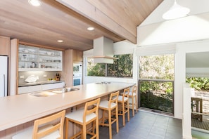 A modern kitchen with a large island, wooden chairs, and a view of greenery outside. - Top Deck (Lorne)
