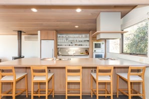 A modern kitchen with a large island, wooden chairs, a built-in oven, and a window with a view of greenery.