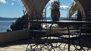 Eine Terrasse im Freien mit Tisch und Stühlen, einem großen Topf mit Pflanze und Blick auf das Meer.