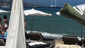 Ein Strand mit Liegestühlen und Sonnenschirmen, ein klarer, blauer Himmel und Segelboote im Hintergrund.