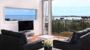 Ein modernes Wohnzimmer mit großem Fernseher, bequemen Sitzgelegenheiten und einem Balkon mit Meerblick.