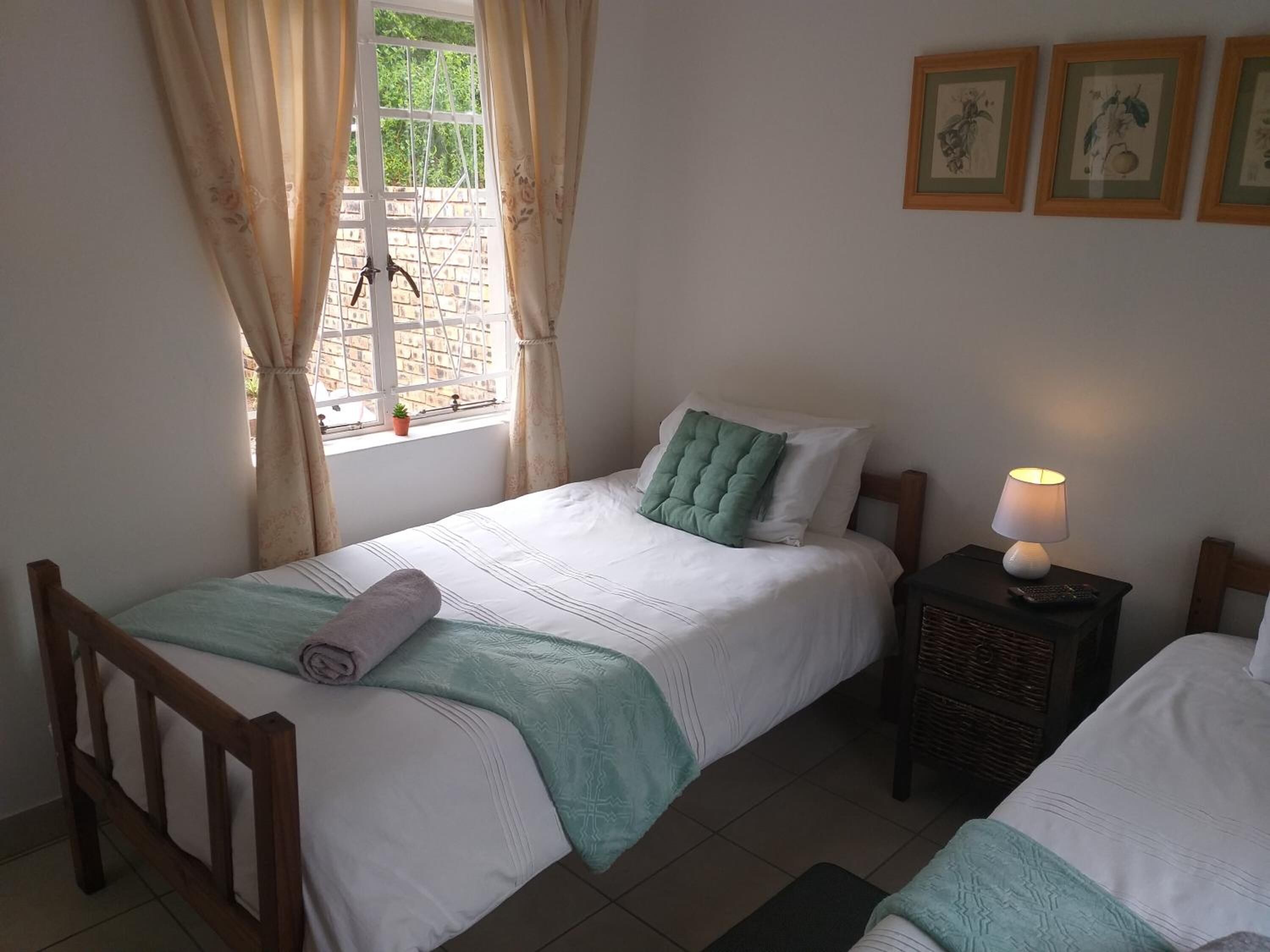 Un dormitorio con dos camas, una ventana con cortinas, una lámpara de noche y cuadros en la pared.