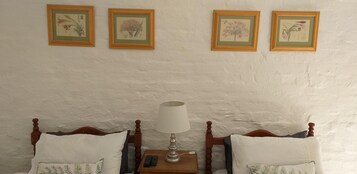 Quarto com duas camas de madeira, uma mesa de cabeceira com abajur, e três impressões botânicas emolduradas na parede.