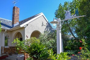 A building with a brick chimney, a white facade, and a garden with various plants and a windmill sculpture. - Padlangs (Kimberley)