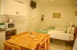 Quarto compacto com uma mesa de jantar de madeira, duas camas simples, micro-ondas e uma pequena cozinha com chaleira.