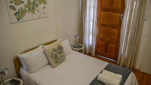 A neatly made bed with white bedding and a grey blanket, a bedside table with a lamp, a painting of flowers on the wall, and a wooden door with curtains.