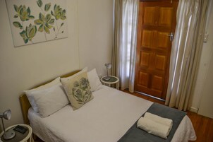 Cama bem arrumada com roupa de cama branca e manta cinza, mesa de cabeceira com abajur, quadro de flores na parede e porta de madeira com cortinas.