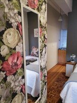 Un dormitorio con un espejo grande, una cama con edredón blanco, piso de madera y una mesita de noche pequeña con una lámpara.
