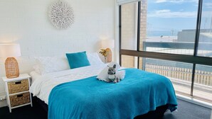 Quarto com uma grande janela, cama com manta azul, roupa de cama branca e um objeto decorativo na parede.