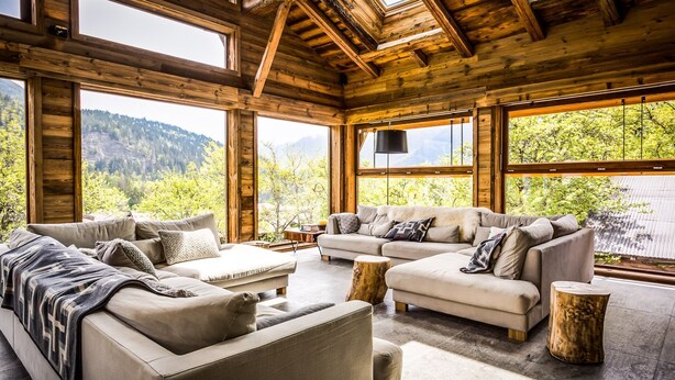 A spacious living room with a large sofa set, a coffee table, and a fireplace. The room features large windows offering a view of a mountainous landscape.