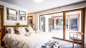 A spacious bedroom with a large bed, a wooden bench, and a chair. There are two mirrors on the wall, and a sliding glass door leads to an outdoor area.