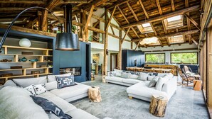 A spacious living area with a high ceiling and exposed wooden beams, featuring a large sofa set, a dining area, and a fireplace.