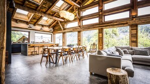 A spacious room with a high ceiling, wooden beams, a dining area with chairs and a table, a sofa, and a view of a forest through large windows.