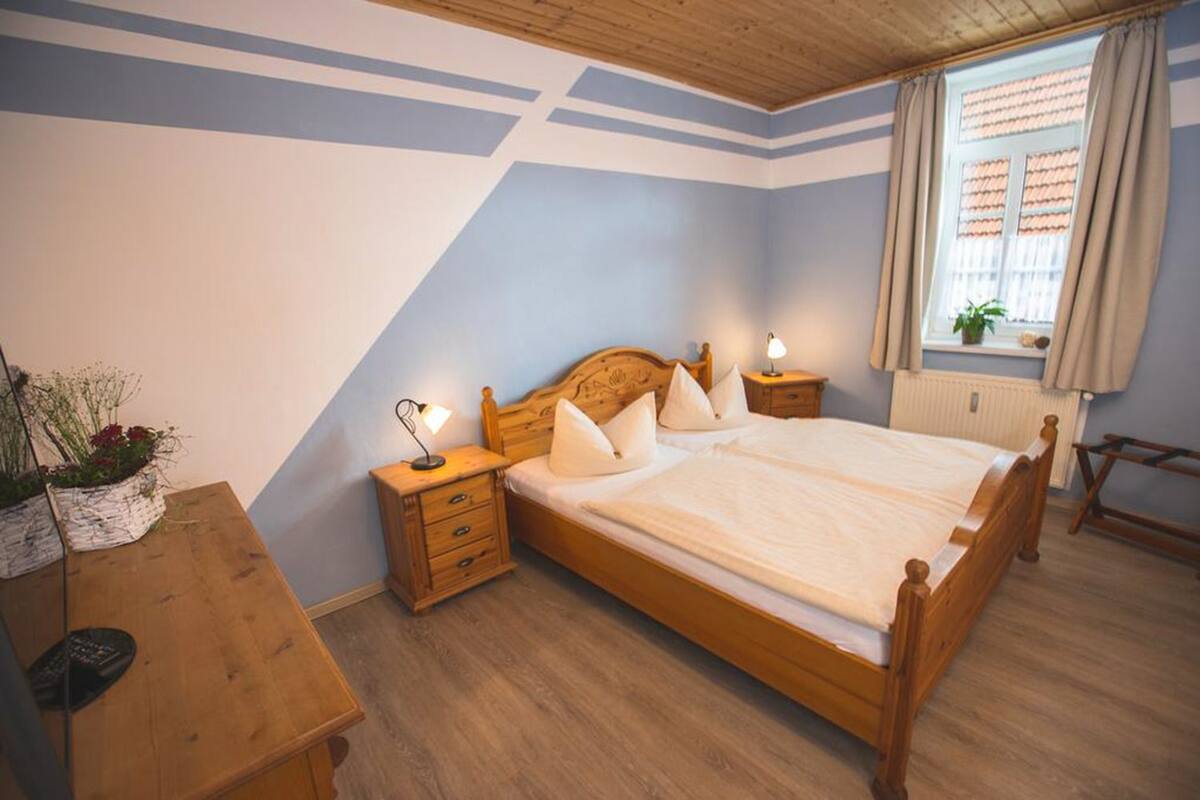 Un dormitorio con cama de madera, mesitas de noche, una planta en el alféizar y cortinas en la ventana.
