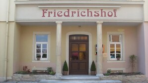 A building with a sign reading "Friedrichshof" above a dark brown door, flanked by two windows and potted plants. - Friedrichshof Restaurant & Pension (Bad Klosterlausnitz)