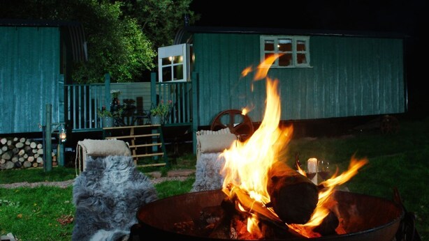 A metal fire pit with flames, surrounded by a grassy area and a wooden deck.