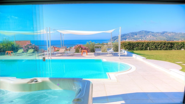 Una piscina con sillones de descanso y un jacuzzi, rodeada por una terraza con vistas al mar y las colinas.