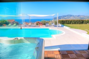 Une piscine dotée de chaises longues et d’un jacuzzi, entourée d’une terrasse offrant une vue sur la mer et les collines.
