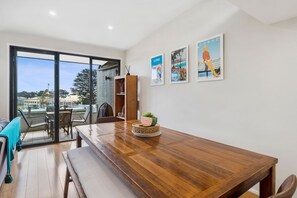 A dining area with a wooden table, chairs, and a bench. There is a bookshelf and a window with a view of the outside. - Sandy Beach Apt 6 (Lorne)
