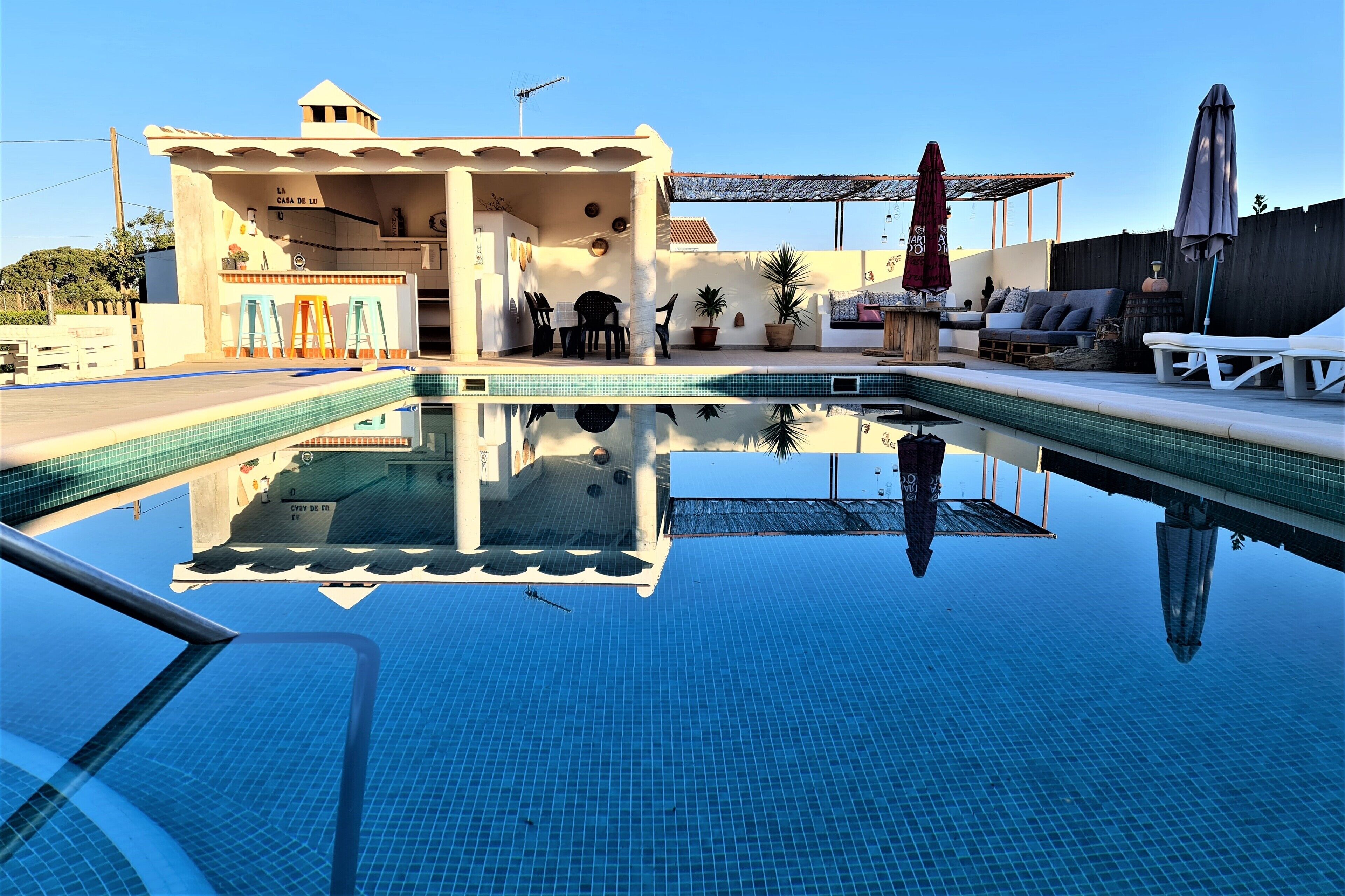 A swimming pool with a clear reflection, a covered seating area with outdoor furniture, and a building with a sign reading "LA CASA DE LU".