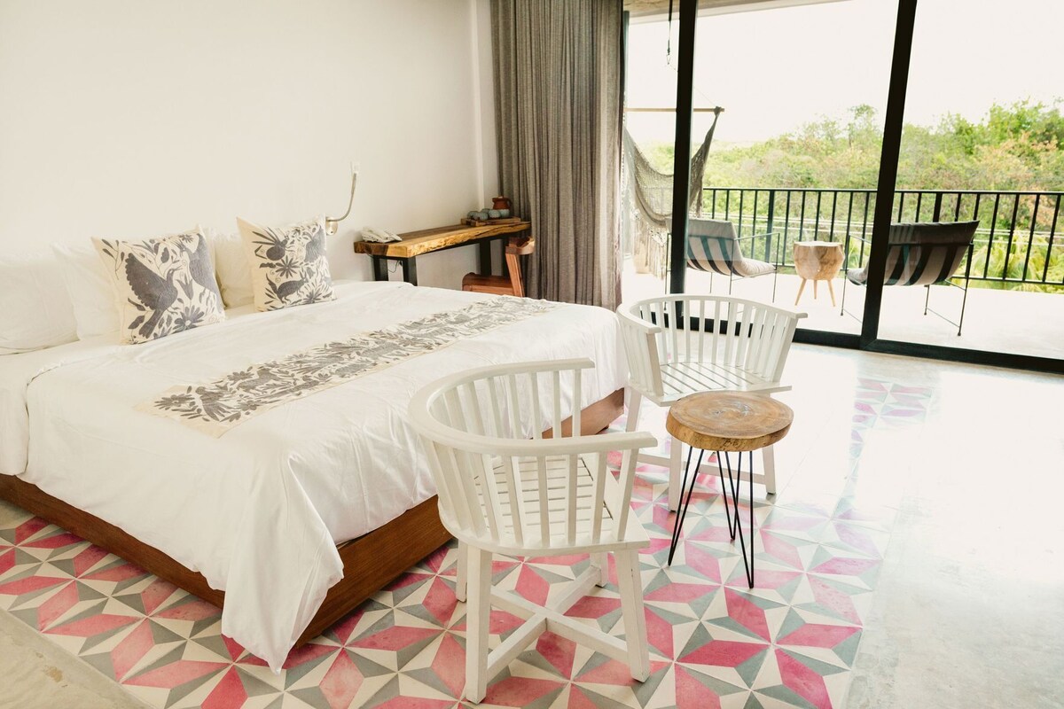 Habitación de hotel con cama, sillas blancas, una mesita redonda pequeña, una mesita de noche de madera y un balcón con mobiliario exterior.