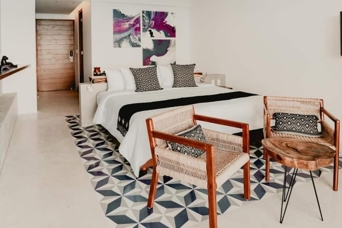 A bedroom with a bed, two wicker chairs, a wooden side table, and a geometric-patterned rug.
