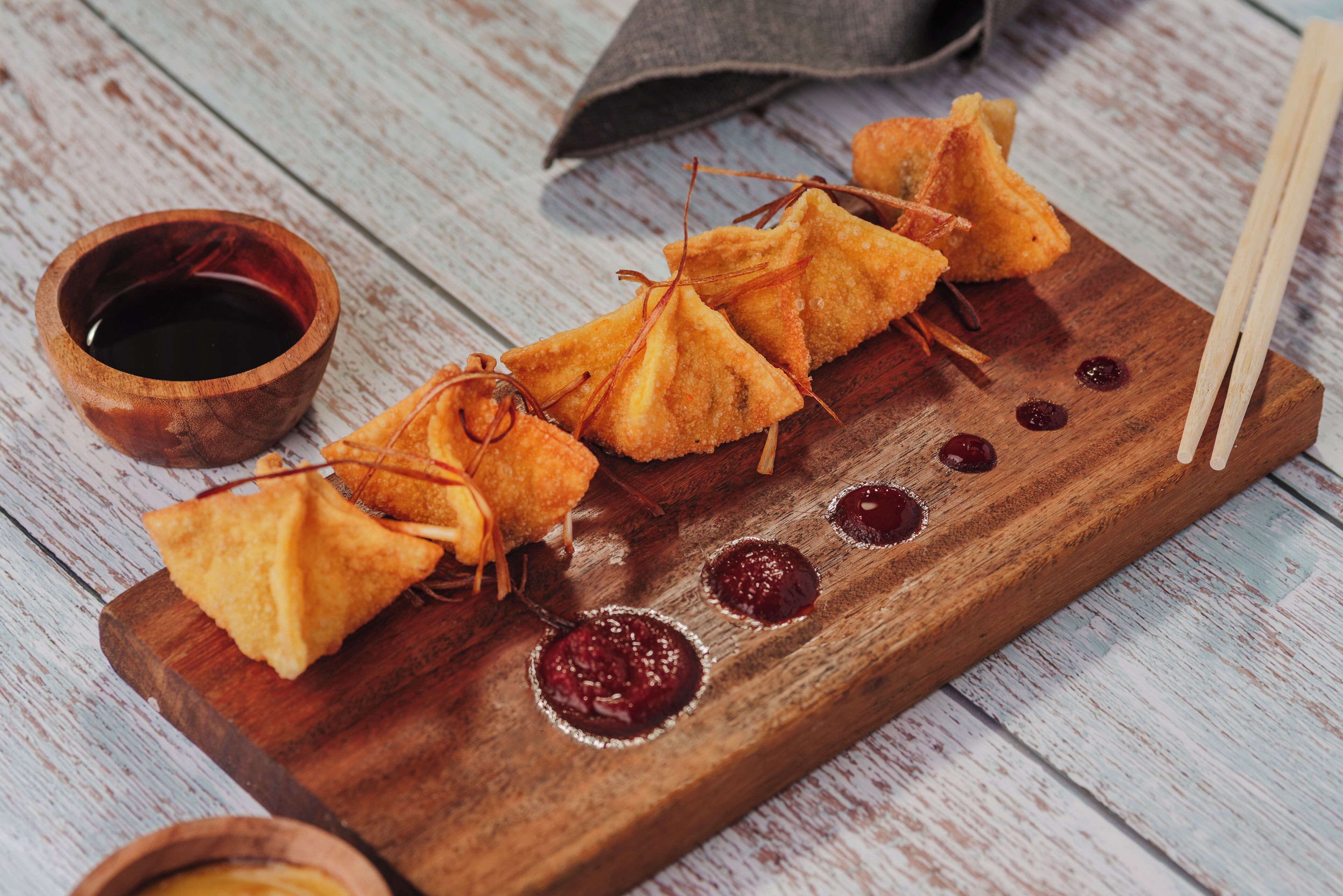 A wooden board with triangular food items, a small bowl of dark liquid, and a pair of chopsticks.