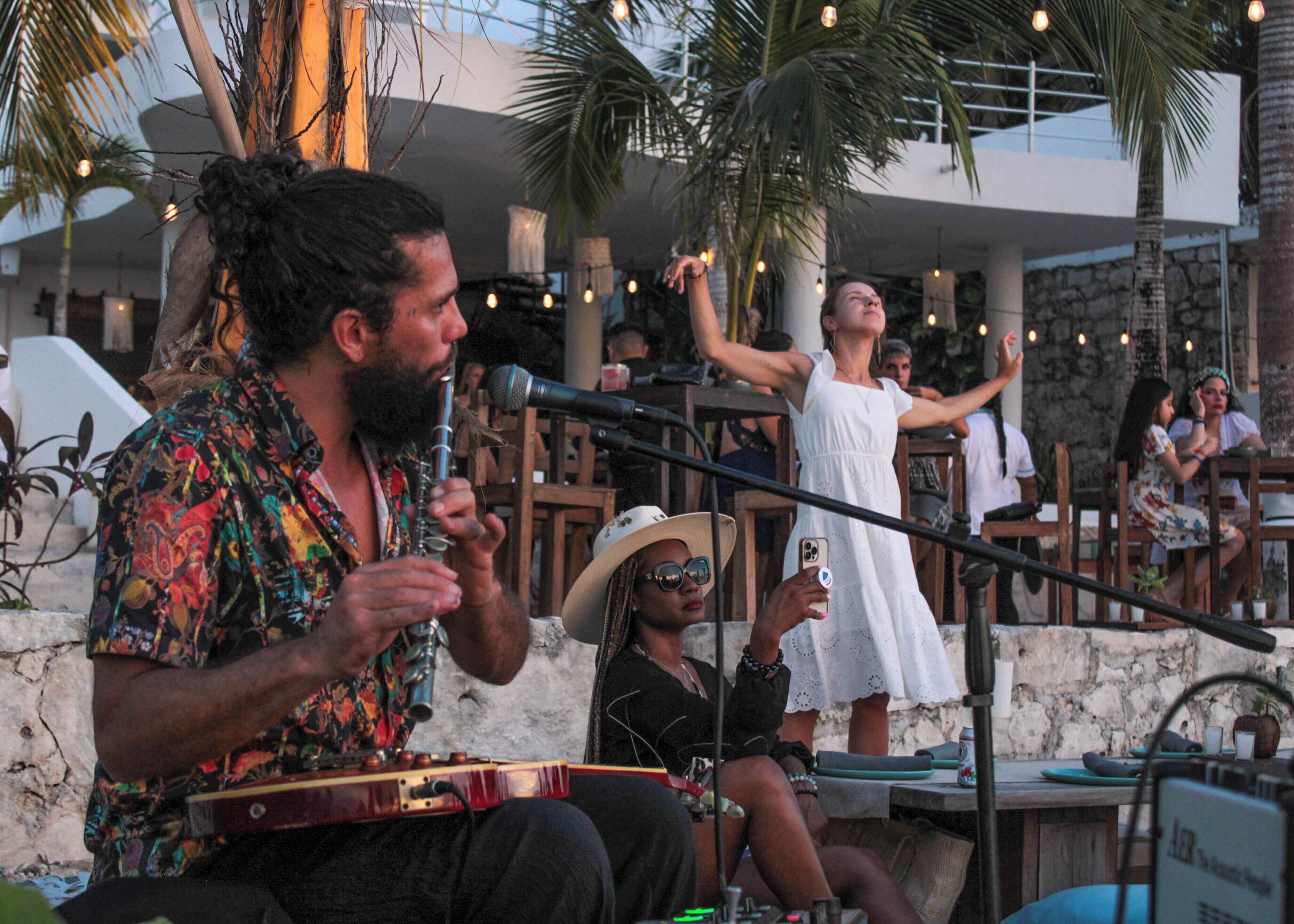 A musician playing a guitar, seated at a table with a microphone. There are palm trees and a stone wall in the background.