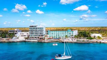 Un hotel frente al mar con varias construcciones, un puerto deportivo con un velero y un cielo azul despejado.