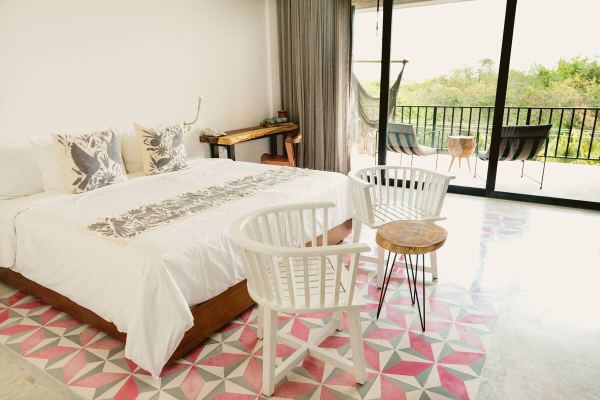 A bedroom with a bed, white chairs, a small table, and a balcony with chairs.