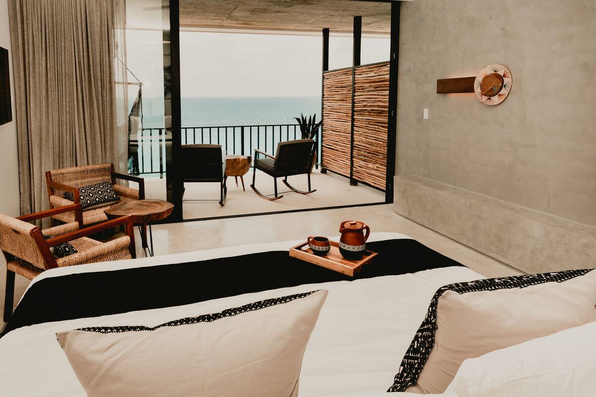 A hotel room with a bed, two rocking chairs, a small table with a tray of drinks, and a view of the ocean through large windows.