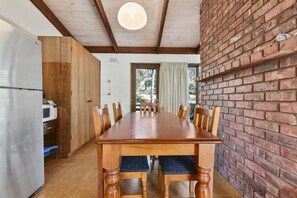 A dining area with a wooden table and chairs, a brick wall, and a view through a window. - Aireys 15 (Aireys Inlet)
