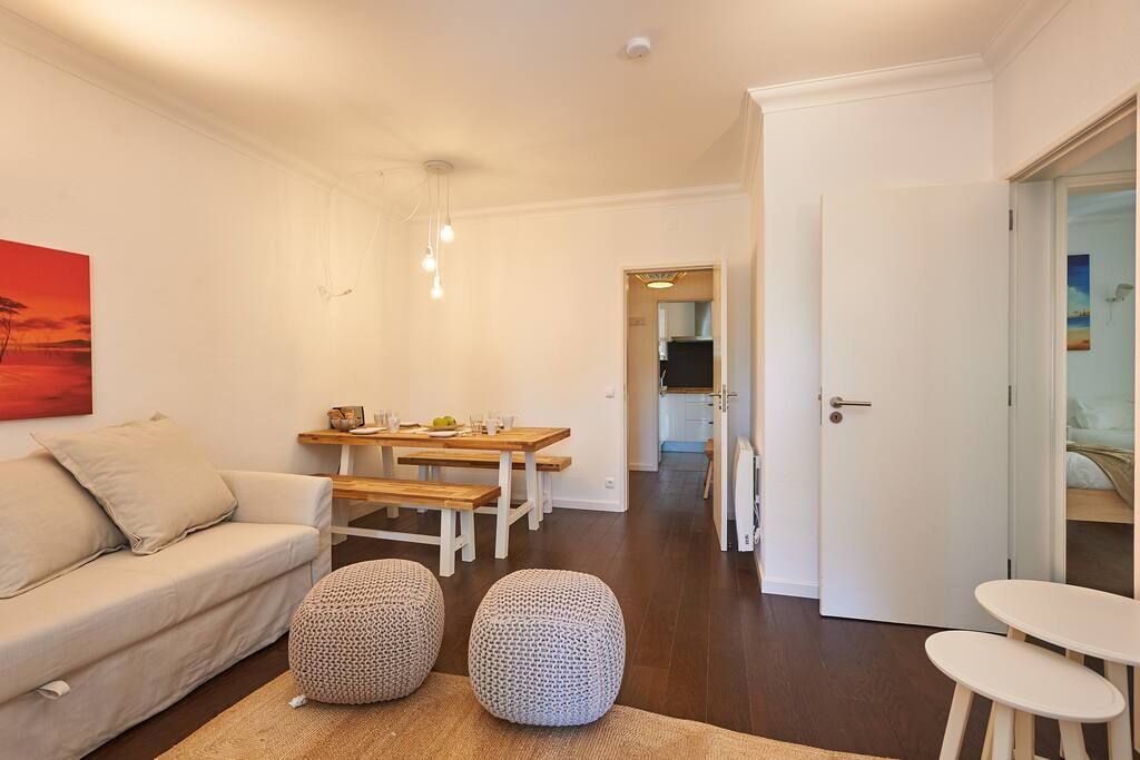 Una sala moderna con un sofá, una mesa de comedor con bancos y dos otomanas redondas. Hay una puerta que da a otra habitación.