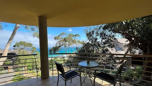 Un balcone con tavolo e sedie, affacciato su una zona costiera con alberi e cielo sereno.