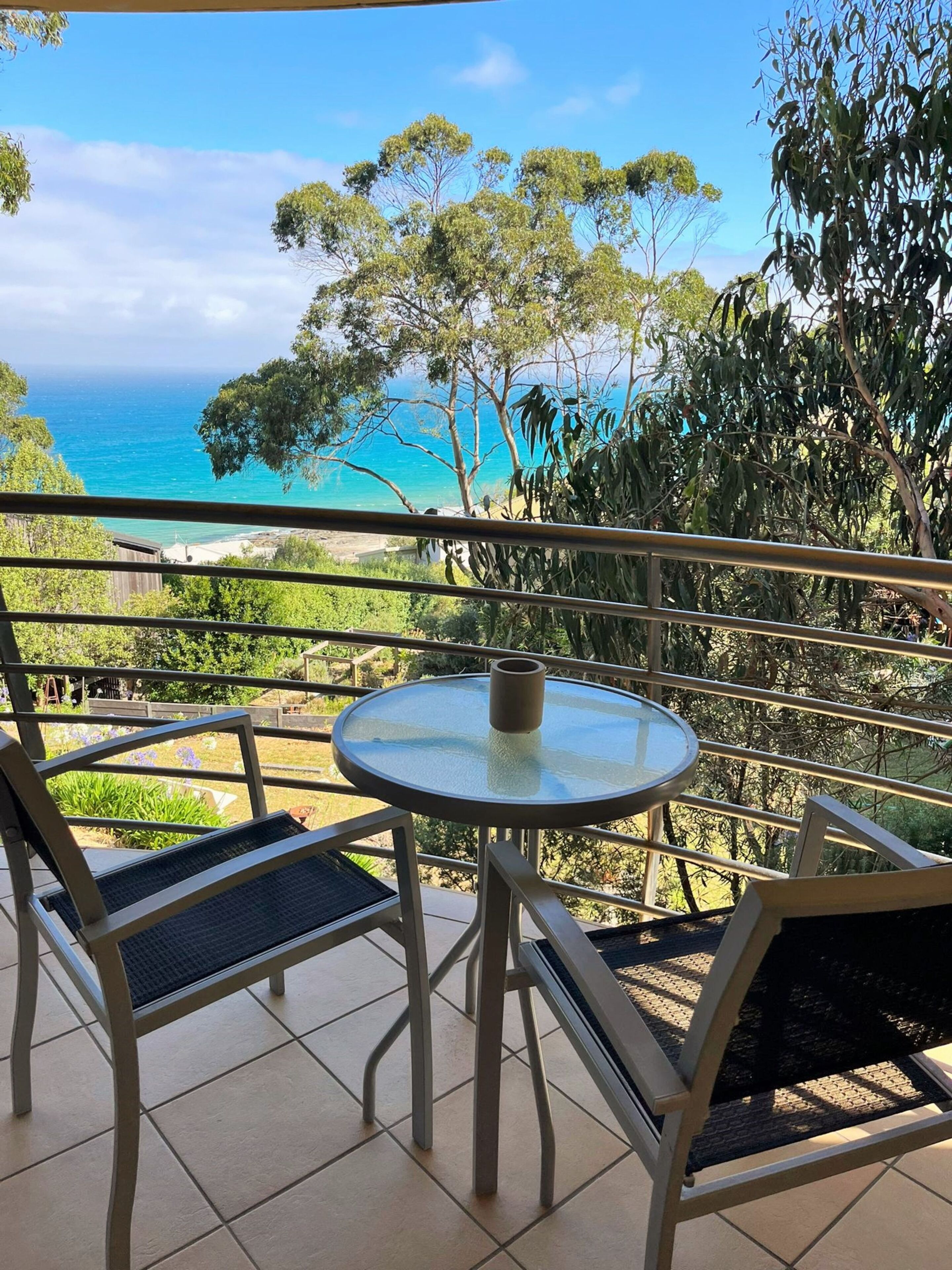 Un balcon avec une table et des chaises en verre, donnant sur un paysage côtier avec des arbres et une vue sur la mer.