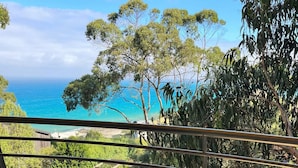 Un balcone con un tavolo in vetro e sedie, affacciato su un paesaggio costiero con alberi e vista sul mare.