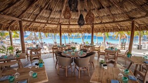 An outdoor dining area with wooden tables and chairs, a thatched roof, and a view of the beach and ocean. - Fully renovated Tropical 3-bdr Villa at Blue Bay Resort, at beach and pool!!! (Blue Bay)