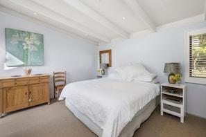 A bedroom with a large bed, a wooden dresser, a chair, a bedside table with a lamp, and a painting on the wall.