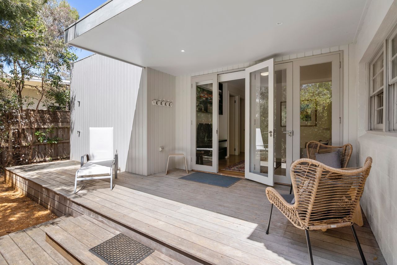 Une terrasse extérieure moderne, dotée d’une plateforme en bois, d’une chaise blanche, d’une chaise en osier et d’une porte vitrée donnant sur un espace intérieur.