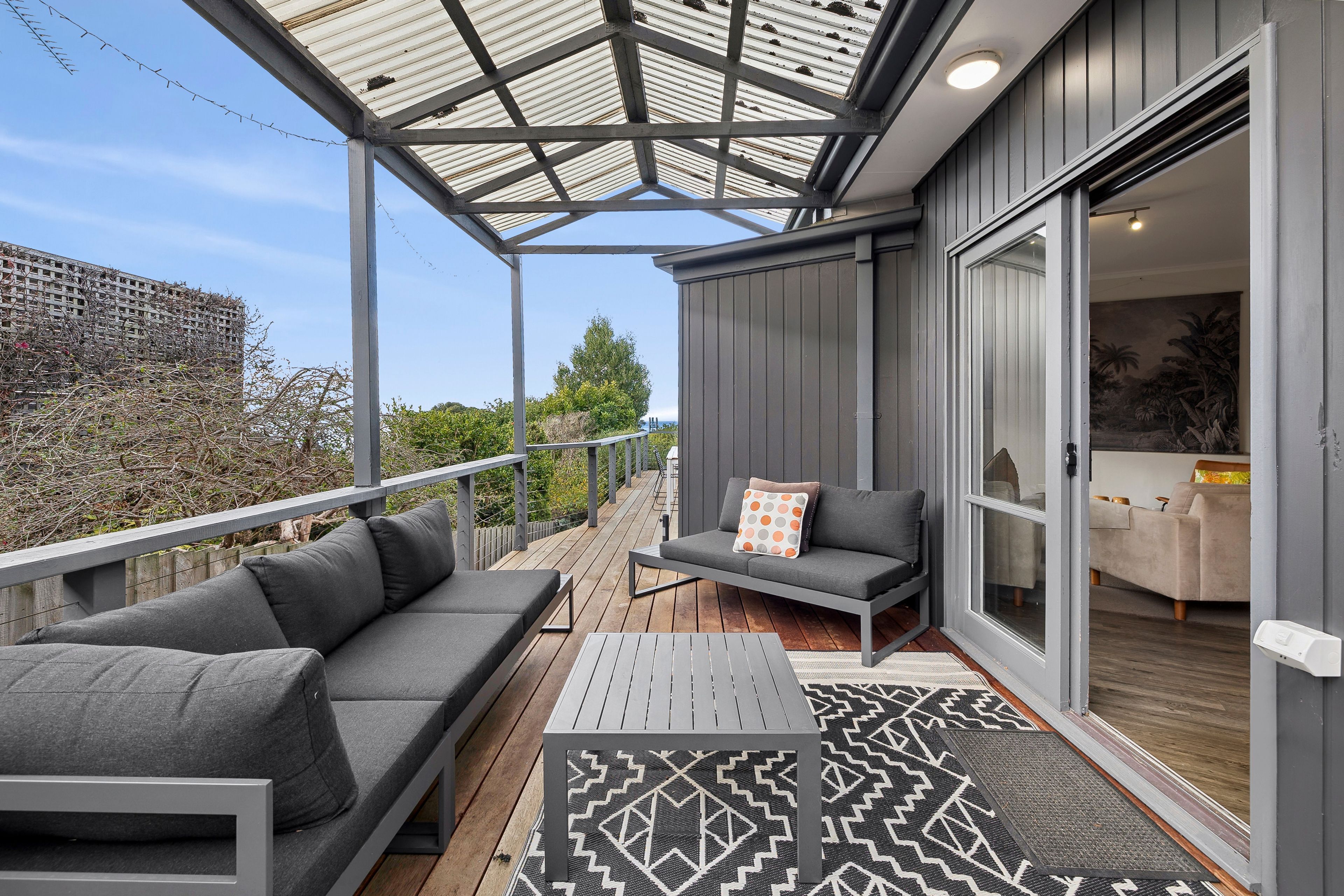 A covered outdoor seating area with a sofa and a table, a sliding glass door leading to a room, and a view of a building in the background.
