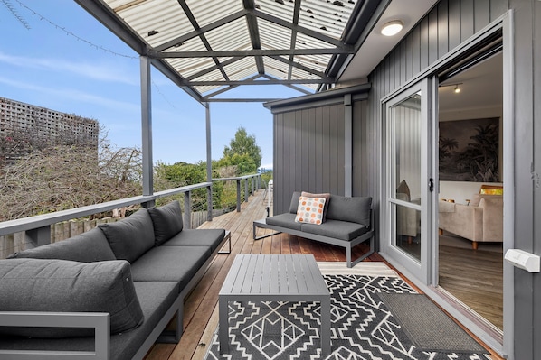 A covered outdoor seating area with a sofa and a table, a sliding glass door leading to a room, and a view of a building in the background.