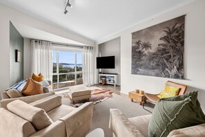 En moderne stue med sofa, sofabord, fjernsyn og et stort vindue med gardiner.