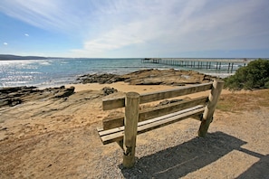 Un banc de fusta a una platja rocosa amb un moll al fons.