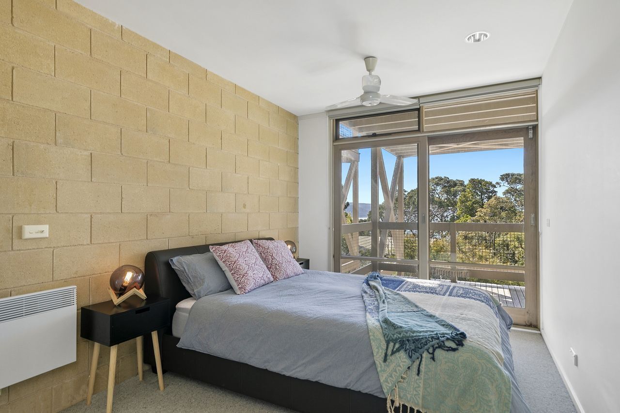 A bedroom with a large bed, a nightstand, a heater, a ceiling fan, and a view of trees through a sliding door.