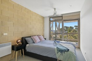 A bedroom with a large bed, a nightstand, a heater, a ceiling fan, and a view of trees through a sliding door.