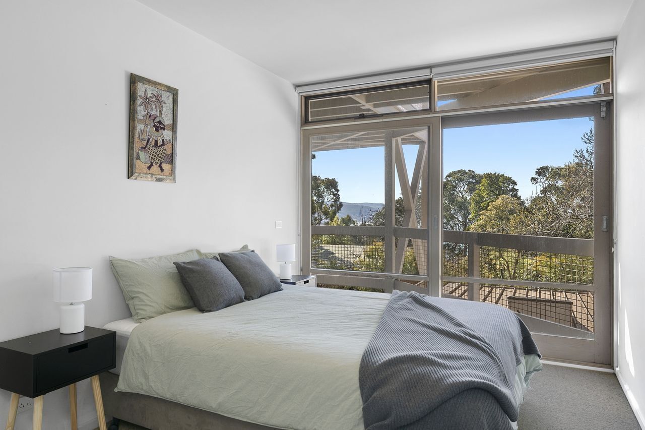 Un dormitorio con una cama grande, una mesita de noche, una lámpara y una ventana con vistas a árboles y una cerca.