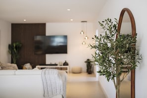 Un salon moderne avec un canapé blanc, un téléviseur à écran plat fixé au mur, un buffet en bois et un grand miroir.